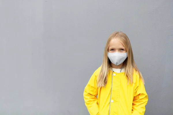 Klein blank meisje 7 jaar oud met blond haar in gele regenjas en gezichtsmasker tijdens Coronavirus pandemie. Kleuren van het jaar 2021 ultieme grijs en verhelderend — Stockfoto