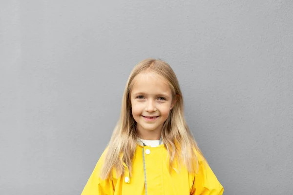 Šťastný malý běloch dívka 6-7 let s blond vlasy ve žlutém plášti stojí venku u šedé zdi. Barvy roku 2021 konečná šedá a osvětlující — Stock fotografie