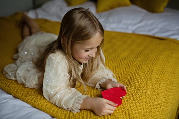 Een schoolmeisje met een rood hart. Valentijnsdag tijdens pandemische coronavirus covid-19 quarantaine. Kind tonen hart online via video chat naar jongen vriend — Stockfoto