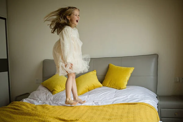 Schöne kleine Mädchen in stilvollem Kleid springen auf dem Bett mit weißer Decke zu Hause. Bleiben Sie zu Hause, während Coronavirus covid-19 Lockdown und haben Sie Spaß. Glückliche Kindheit — Stockfoto