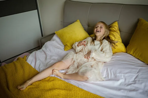 Gyönyörű kislány elegáns ruhában ugrál az ágyon fehér takaróval otthon. Maradj otthon a Coronavirus Covid-19 lezárása alatt és érezd jól magad. Boldog gyermekkori koncepció — Stock Fotó