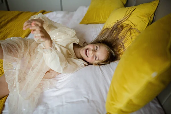 Schöne kleine Mädchen in stilvollem Kleid springen auf dem Bett mit weißer Decke zu Hause. Bleiben Sie zu Hause, während Coronavirus covid-19 Lockdown und haben Sie Spaß. Glückliche Kindheit — Stockfoto
