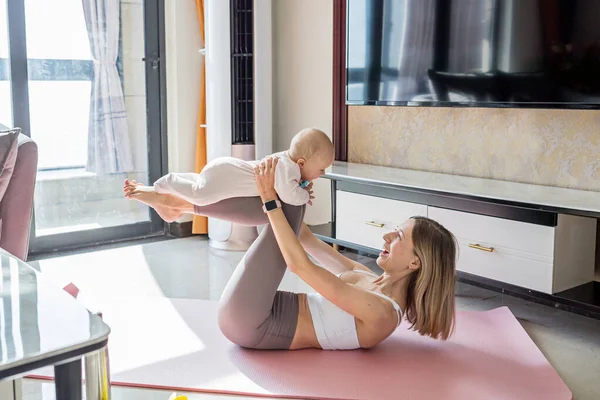 Young mother in sport clothing exercising at home with baby. Online training during coronavirus covid-19 quarantine. Stay fit and safe during pandemic lockdown. Sport, fitness, healthy concept