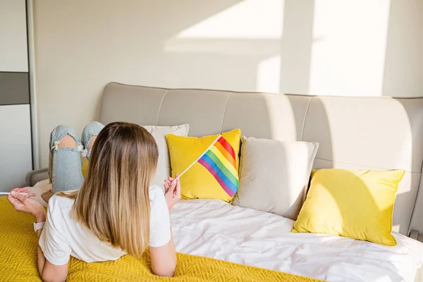 Frau mit Regenbogenfahne auf dem Bett. Lesben feiern Stolz Monat während Covid-19 Coronavirus Pandemie Quarantäne, zu Hause bleiben, soziale Distanzierung und Selbstisolation Konzept — Stockfoto