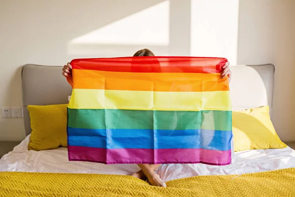 Frau mit Regenbogenfahne auf dem Bett. Lesben feiern Stolz Monat während Covid-19 Coronavirus Pandemie Quarantäne, zu Hause bleiben, soziale Distanzierung und Selbstisolation Konzept — Stockfoto