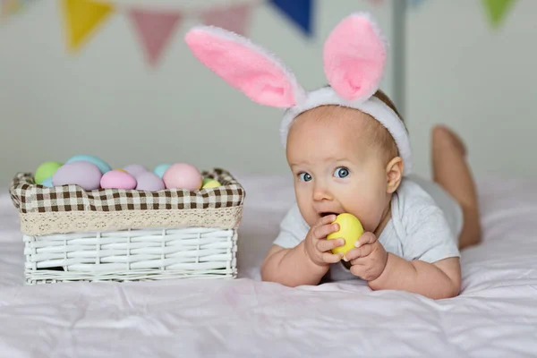 Niña caucásica feliz de seis meses de edad con diadema de orejas de conejo, acostado en la cama en casa en el dormitorio con huevos de Pascua de colores — Foto de Stock