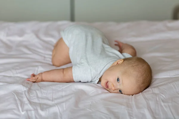 Bebê loiro caucasiano de seis meses deitado na cama em casa. Um miúdo com roupas cinzentas giras. Quarto decorado com bandeiras. — Fotografia de Stock