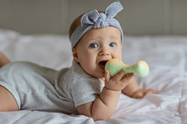 Caucasiano bebê loiro seis meses de idade deitado na cama em casa e brincando com o brinquedo chocalho. Um miúdo com roupas cinzentas giras. Quarto decorado com bandeiras. — Fotografia de Stock