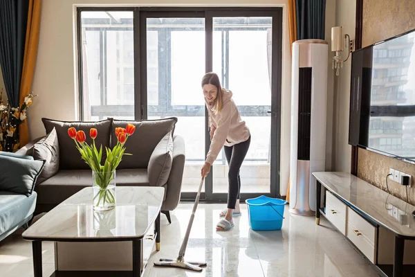 Bella giovane casalinga bionda caucasica che pulisce il pavimento in soggiorno. Interni domestici moderni, vita domestica — Foto Stock