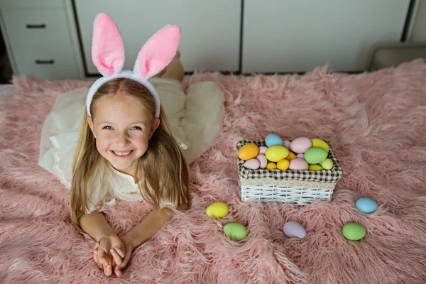 Niña caucásica feliz niño 7 años de edad con orejas de conejo diadema, acostado en la cama en casa en el dormitorio con huevos de Pascua de colores. Permanecer en casa durante la pandemia del Coronavirus covid-19 — Foto de Stock