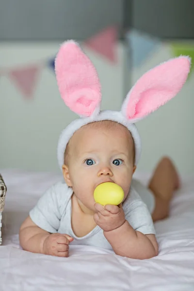 Niña caucásica feliz de seis meses de edad con diadema de orejas de conejo, acostado en la cama en casa en el dormitorio con huevos de Pascua de colores — Foto de Stock