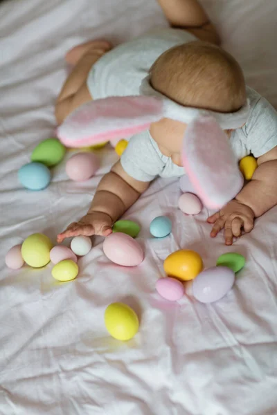 Niña caucásica feliz de seis meses de edad con diadema de orejas de conejo, acostado en la cama en casa en el dormitorio con huevos de Pascua de colores — Foto de Stock