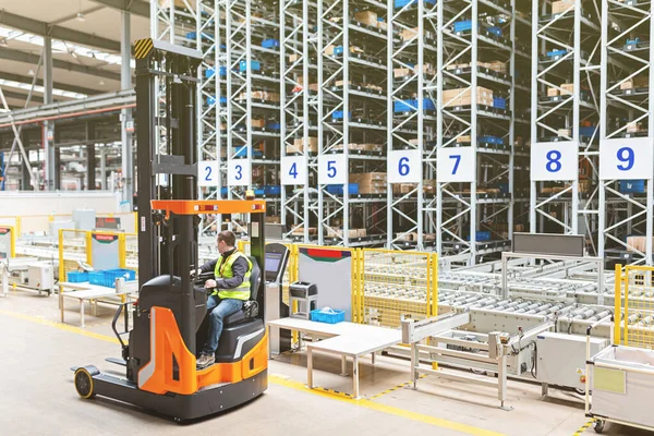 Storehouse employee in uniform working on forklift in modern automatic warehouse. Boxes are on the shelves of the warehouse. Warehousing, machinery concept. Logistics in stock.