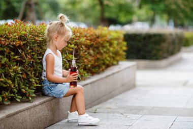 Şirin, şık, sarı saçlı, sıcak yaz gününde açık havada soda içen çocuklar için sağlıksız bir içecek.