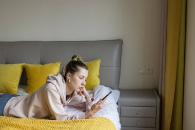 Kafkasyalı genç bir kadın cep telefonu ve kablosuz kulaklık kullanıyor, dinleniyor, yatakta oturuyor ve podcast ya da kulüp binasını dinliyor - sadece sesli sosyal medya uygulaması, sesli sohbet