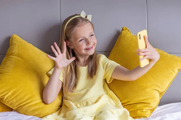 Carino piccola ragazza caucasica con i capelli biondi in abito alla moda illuminante colore giallo seduto a casa durante la quarantena pandemica coronavirus e utilizzando il telefono cellulare. Rimanere a casa durante covid-19 — Foto Stock