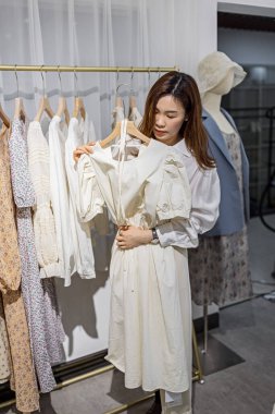 Modern mağazasında kadın giyim kuşamlarıyla çalışan mutlu Asyalı kadın girişimci portresi, genç Çinli kadın sahibi ya da konyak dükkanında dururken elinde şık kıyafetler tutan danışman.