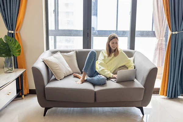 Young beautiful woman wearing yellow hoodie and resting on gray textile sofa at home. Attractive slim female in domestic situation, staying home, relaxing on couch in her apartment. Background, copy