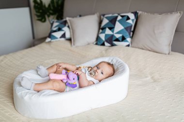 Evde kozanın içinde yatan sevimli sarışın bebek üç aylık. Çocuk doldurulmuş ses oyuncağını tutuyor, meme ucunu emiyor.. 