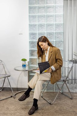 Genç ve güzel stil sahibi Asyalı kadın sandalyede oturup dergi okuyor ve modern ofis ya da kafenin gevşemesini sağlıyor.. 