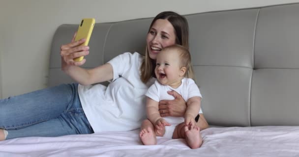 Glad kaukasiska mor och hennes dotter har online videosamtal med far eller mormor via mobiltelefon när du ligger på sängen i mysiga sovrum hemma. Långsamma rörelser — Stockvideo