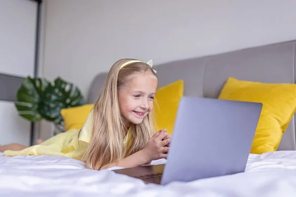 Carino piccola ragazza caucasica con i capelli biondi in abito alla moda illuminante colore giallo seduto a casa durante la quarantena pandemica coronavirus e utilizzando il computer portatile. Rimanere a casa durante covid-19 — Foto Stock