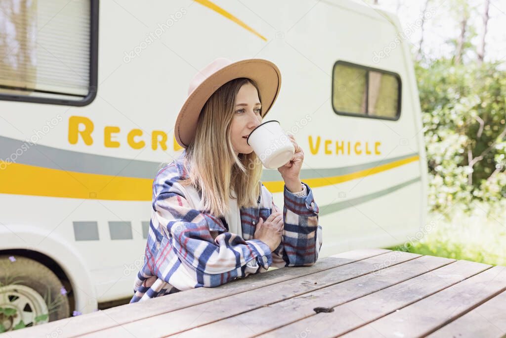 Beautiful millennial caucasian woman posing near camper van in forest. Young stylish woman in shirt and cowboy hat enjoying life and having fun on summer vacation