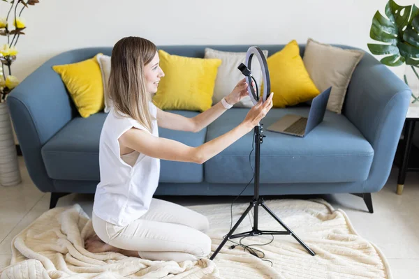 Jovem mulher fazendo conteúdo de foto ou vídeo para mídias sociais com smartphone e luz da lâmpada do anel. Beleza blogueiro sorri para a tela do telefone móvel. Influenciador Faz transmissão se comunica com seguidores — Fotografia de Stock