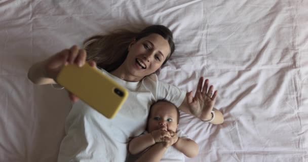 Glückliche kaukasische Mutter und ihre kleine Tochter telefonieren online mit Vater oder Großmutter per Handy, wenn sie zu Hause im gemütlichen Schlafzimmer auf dem Bett liegen. Zeitlupe — Stockvideo