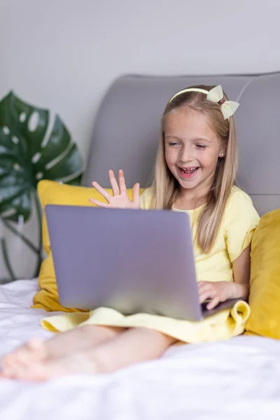 Милая маленькая кавказская девочка с светлыми волосами в модном платье, сидящая дома в светло-желтом цвете во время пандемии коронавируса и используя ноутбук. Оставайтесь дома во время covid-19 — стоковое фото