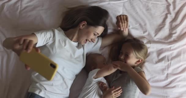 幸せな白人の母親と子供たちは、自宅の居心地の良いベッドルームでベッドの上に横たわっているときに携帯電話を介して父または祖母とオンラインビデオ通話をしています。スローモーション — ストック動画
