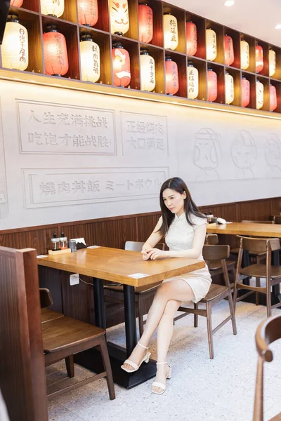 Schöne junge asiatische Frau in traditioneller weißer Kleidung namens Cheongsam sitzt im japanischen Restaurant oder Cafeteria und wartet auf bestelltes Essen. Interieur des Restaurants mit bunten Papierlaternen dekoriert — Stockfoto