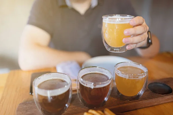 Homem variedade de amostragem de cerveja artesanal sazonal no pub. Amostradores de cerveja em pequenos copos colocados individualmente em furos formados em bandeja de madeira única. Foco seletivo com profundidade de campo rasa. — Fotografia de Stock