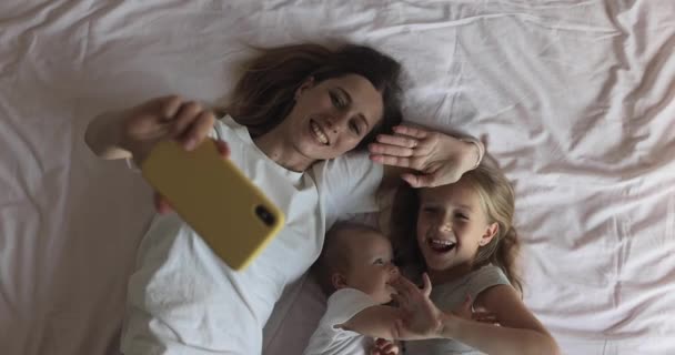 Feliz madre caucásica y los niños tienen videollamada en línea con el padre o la abuela a través del teléfono móvil al acostarse en la cama en el acogedor dormitorio en casa. Movimiento lento — Vídeo de stock