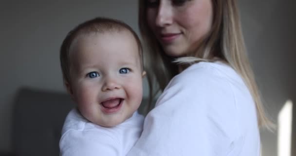 Jeune mère caucasienne et sa petite fille embrassant et jouant à la maison dans la chambre. Petit enfant de sept mois passer du temps avec maman bras à la maison près de la fenêtre, au ralenti. — Video