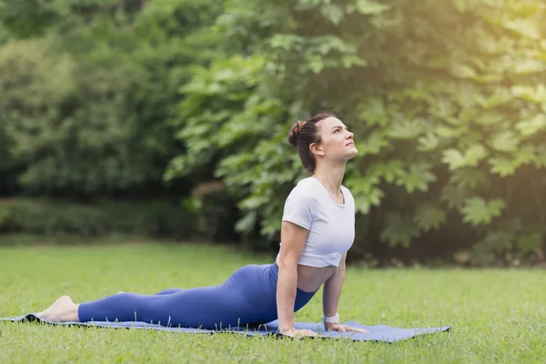 Porträtt av glad ung kaukasisk kvinna som utövar yoga utomhus tidigt på morgonen. Vacker flicka tränar asana på blå yogamatta på grönt gräs i parken på sommaren — Stockfoto