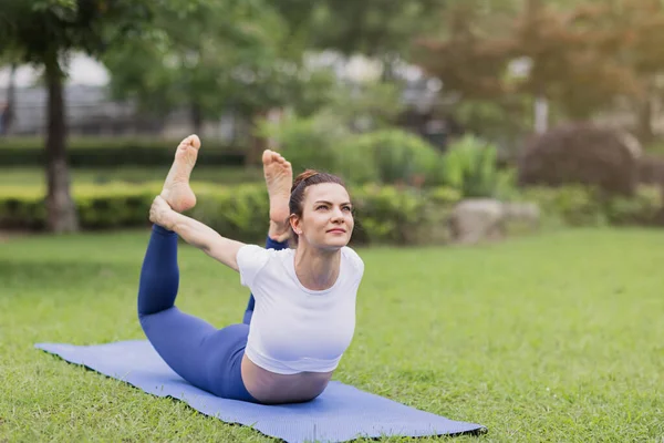 Porträtt av glad ung kaukasisk kvinna som utövar yoga utomhus tidigt på morgonen. Vacker flicka tränar asana på blå yogamatta på grönt gräs i parken på sommaren — Stockfoto