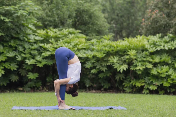 Porträtt av glad ung kaukasisk kvinna som utövar yoga utomhus tidigt på morgonen. Vacker flicka tränar asana på blå yogamatta på grönt gräs i parken på sommaren — Stockfoto