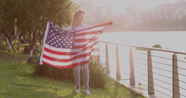 Chica ondeando bandera nacional de EE.UU. al aire libre. Mujer joven con cabello rubio en ropa casual bailando en el parque al atardecer con bandera americana. Feliz día de independencia 4 de julio. Movimiento lento — Vídeos de Stock