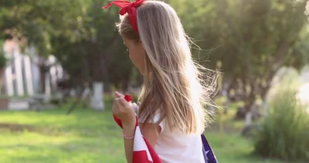 4k. Kölyök lengeti a nemzeti amerikai zászlót a szabadban. Aranyos kislány hét éves szőke hajjal alkalmi ruhában, parkol a naplementében amerikai zászlóval. Boldog függetlenség napját július 4-én. Lassú. — Stock videók