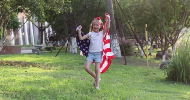 4k. Kölyök lengeti a nemzeti amerikai zászlót a szabadban. Aranyos kislány hét éves szőke hajjal alkalmi ruhában táncol a parkban naplementekor amerikai zászlóval. Boldog függetlenség napját július 4-én. Lassú. — Stock videók