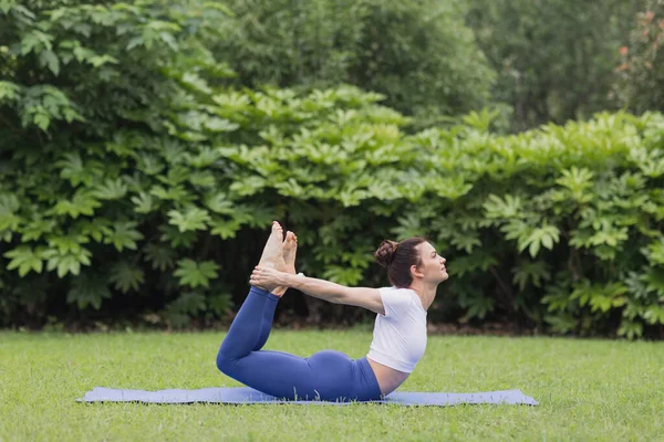 Porträtt av glad ung kaukasisk kvinna som utövar yoga utomhus tidigt på morgonen. Vacker flicka tränar asana på blå yogamatta på grönt gräs i parken på sommaren — Stockfoto