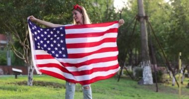 Ulusal Amerikan bayrağını sallayan bir kız. Sarı saçlı, günbatımında Amerikan bayrağıyla parkta dans eden genç bir kadın. Bağımsızlık günün kutlu olsun 4 Temmuz. Yavaş çekim