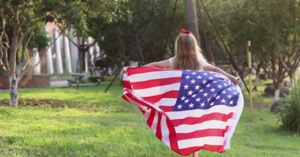 4k. Kölyök lengeti a nemzeti amerikai zászlót a szabadban. Aranyos kislány hét éves szőke hajjal alkalmi ruhában, parkol a naplementében amerikai zászlóval. Boldog függetlenség napját július 4-én. Lassú. — Stock videók