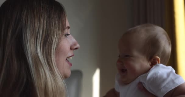 Giovane madre caucasica e la sua bambina abbracciano e giocano a casa in camera da letto. Bambino di sette mesi che trascorre del tempo con le braccia della mamma a casa vicino alla finestra, al rallentatore. — Video Stock