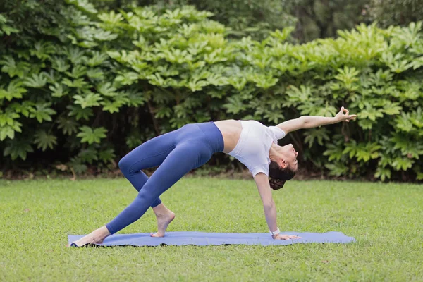 Porträtt av glad ung kaukasisk kvinna som utövar yoga utomhus tidigt på morgonen. Vacker flicka tränar asana på blå yogamatta på grönt gräs i parken på sommaren — Stockfoto