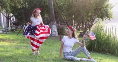 Sevimli küçük kız yedi yaşında ve sarı saçlı genç anne günbatımında parkta ulusal Amerikan bayrağını sallıyor. Bağımsızlık günün kutlu olsun 4 Temmuz. Yavaş çekim
