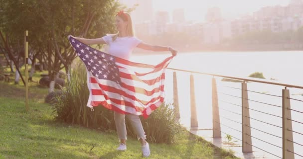 Fille agitant drapeau national des États-Unis à l'extérieur. Jeune femme aux cheveux blonds en tenue décontractée dansant dans le parc au coucher du soleil avec drapeau américain. Joyeux jour de l'indépendance le 4 juillet. Mouvement lent — Video