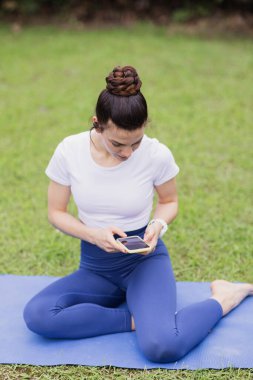 CoVID-19 Coronavirus salgını sırasında yaz parkında sanal yoga dersinden sonra dinlenen beyaz kadın. Fitness egzersizi yaparken sosyal mesafeler. Cep telefonu tutan, müzik dinleyen bir kız