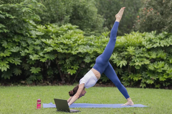 Ung kaukasisk kvinna tränar online i sommarparken under COVID-19 coronavirus pandemi. Social distansering under konditionsträning. Flicka som bär sportkläder när du stretchar och utövar yoga — Stockfoto